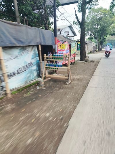 The image shows a street scene in Baleendah, Indonesia. A person is riding a motorcycle on the paved road. The motorcycle is traveling away from the camera. On the right side of the road is a sidewalk with a small wooden structure with bottles on shelves. The wooden structure is in front of a small building with a sign that says "Soda". There is a large, dark blue tarp-like covering on the left side of the road. There is foliage on the right side of the road and trees in the background.