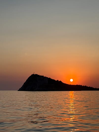 The image depicts a sunset over the sea with a silhouetted island in the foreground, located in Mirišta, Montenegro. The sky transitions from a pale blue at the top to a warm orange hue near the horizon, where the sun is prominently visible as a bright, circular orb. The island, which appears to be a fortified structure or a rocky outcrop with some buildings or ruins on its summit, is cast in shadow against the vibrant sky. The calm sea reflects the colors of the sunset, creating a shimmering path of light from the sun towards the viewer. The overall scene is serene and captures the natural beauty of the coastal landscape at dusk.