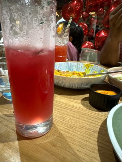 An indoor dining scene is depicted, likely within a restaurant or cafe located in Kalyan-Dombivli, India. In the immediate foreground, a tall, clear glass contains a red, frothy beverage with ice cubes, resting on a light-colored wooden table. To the right, a striped ceramic bowl holds a partially consumed yellow dish, with a metal fork present within it. Adjacent to this, a small black ramekin contains an orange-colored sauce. A clear glass with blue liquid is partially visible on the far left. The background is out of focus, revealing numerous reflective red heart-shaped balloons suspended in the air, indicating a celebratory or themed ambiance. Partially visible figures include a person with dark hair wearing a pink top, seen from behind, and an adult's hand on the far right. Another glass containing an orange drink, garnished with an orange slice and a green and white striped straw, is also present in the mid-background.
