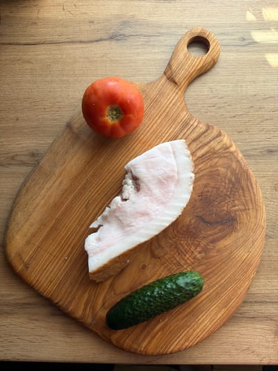This is an overhead shot of a rustic wooden cutting board positioned on a wooden table. The cutting board is oval-shaped with a handle featuring a circular hole. Placed on the board are three food items: a small, bright red tomato, a thick slice of raw pork fat with streaks of pink and brown, and a whole, bumpy green cucumber. The lighting suggests it is daytime, possibly late morning or early afternoon, with sunlight casting faint shadows from unseen objects onto the table surface to the left of the cutting board. The overall scene is simple, focusing on the raw ingredients, and evokes a sense of preparation or a light meal. There is no visible text in the image. The location context is Uhersko, Ukraine.