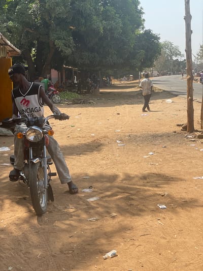 An outdoor scene in Gidan Kado, Nigeria, depicts a daytime market or roadside area. In the foreground, a male individual is seated on a red and chrome motorcycle with his feet on the reddish-brown dirt ground. He wears a black beanie, a white mesh sleeveless shirt with "BALENCIAGA PARIS" in black lettering, and light-colored trousers with dark sandals. His hands are on the motorcycle handlebars. The ground is unpaved, scattered with plastic debris and litter, with tree shadows cast upon it.
In the midground, to the right, a second male individual wearing a light-colored, patterned short-sleeved shirt and dark pants walks away from the camera on the dirt shoulder adjacent to an asphalt road. Two bare wooden poles stand vertically near the road's edge.
To the left and center midground, beneath a large, leafy green tree, is an active market area. Multiple individuals are visible, some standing and others seated. Piles of light-green produce are arranged on the ground next to a red and silver motorcycle parked near simple, partially visible structures with corrugated roofs. The asphalt road extends into the background, where more people and vehicles are discernible under a bright, clear sky.