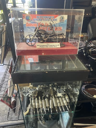 The image depicts an indoor display area within the city of Ban Yid, Thailand, showcasing motorcycle memorabilia and related items.

A multi-tiered display stand, approximately 1.5 meters in height, occupies the central frame. The uppermost section features a clear acrylic case, measuring approximately 0.5 meters in width, 0.3 meters in height, and 0.2 meters in depth. Inside this case, a miniature metal motorcycle model, approximately 0.2 meters long, rests on a red, velvet-covered platform. Behind the model, a sign is visible with text "BUTTERWORTH BIKERS CLUB @ BBC Convoy Ride To PHUKET BIKE WEEK 2011" and associated logos. A smaller plaque in front of the model reiterates "BUTTERWORTH BIKERS CLUB Phuket bike week 2011".

Below the top display, a second glass-enclosed section of the stand, framed in black, contains obscured items including what appear to be printed materials or magazines. Adjacent to the right of this section, a black motorcycle seat with a studded black backrest and a black metal frame are visible, indicative of a full-sized motorcycle or a dedicated display.

The lowest section of the display stand is also a glass-enclosed case, exhibiting various objects. Approximately 10-12 silver-colored, cylindrical items, identified as tattoo needle cartridges or grips with pointed tips, are arranged in rows. Some bear numerical labels such as "000004" and "000005". To the left of these, a small, decorative, antique-style revolver model with a light-colored grip is horizontally positioned. Other miscellaneous metal components, possibly keychains or small parts, are also present within this case.

To the far left of the display stand, a black wire rack holds several magazines or brochures. The environment is illuminated by bright natural light entering from the left, causing reflections on the glass surfaces of the display cases. The floor appears to be concrete. The overall setting suggests a specialized retail establishment, a club facility, or a themed exhibit space.