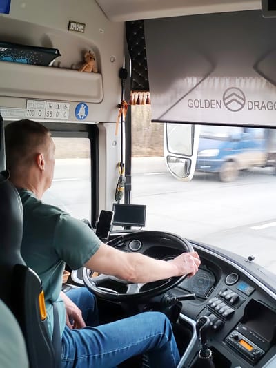 The image captures the interior of a bus cockpit from a passenger's perspective, looking forward. A male bus driver, seen from behind, is wearing a green t-shirt and blue jeans, with his hands on the steering wheel. The bus appears to be in motion on a multi-lane highway, with the outside view showing a blurred passing blue vehicle (likely a truck or another bus) through the windshield and side mirror.
The bus cabin is well-equipped with a detailed dashboard featuring a manual gear stick, various control buttons, a radio/cassette player, and digital displays. A mobile phone is mounted in a holder, and a small monitor screen is visible to the driver's right. Overhead, a beige compartment holds a small light brown teddy bear and a miniature framed picture containing three portraits. A dark blue tablet is also stored in this compartment.
A grey sunshade is partially pulled down, displaying the text "GOLDEN DRAGON" and a stylized logo. Near the side window, a decorative curtain with tassels and a St. George's ribbon are visible. Above the driver, on an interior panel, there are numbers and icons: "700 0 55 0 0", "КРЫМ 0" (Krym 0), and several pictograms indicating standing capacity, elderly/disabled access, and a blue circular icon depicting a person with a child.
The time of day appears to be daytime, with a clear, though possibly overcast, sky. The driver appears focused on the road, suggesting a routine journey. The presence of the St. George's ribbon aligns with the given location context of Krasnyi Bor, Russia, and is a common cultural accessory in that region.