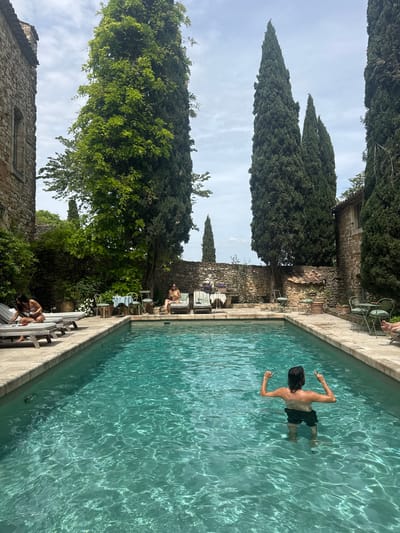 The image depicts a rectangular swimming pool in Saint-Victor-la-Coste, France. A man with long dark hair and black swim shorts is in the pool with his arms raised. Two people are sitting on lounge chairs along the pool's edge. A stone wall borders the area with tall, narrow trees interspersed along the perimeter. Other chairs and tables are present on the poolside.