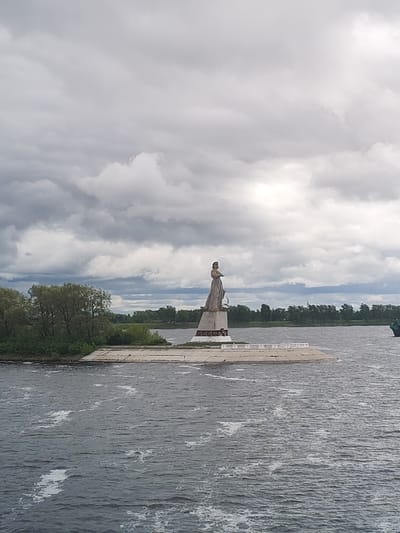 The image depicts a statue situated on an artificial island within the Volga River in Rybinsk, Russia. The statue, a prominent feature, is situated atop a decorated pedestal. The artificial island features a concrete foundation with a white railing around the perimeter. The Volga River is visible in the foreground. A forested area lines the bank on the left side of the image, and a few buildings are visible in the distance. The sky is overcast with dense, grey clouds.