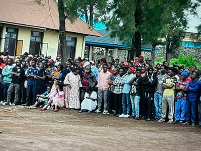 A large group of people, mostly men, are gathered in front of a building in Keshero, Democratic Republic of the Congo. The building is a one-story structure with a yellow facade and a tiled roof. It has a sign above the door. The people are standing in a line, facing the building. They are all wearing casual clothing. There are several trees in the background. The ground is dirt. It appears the people are waiting to enter the building. It is unclear what their purpose is.