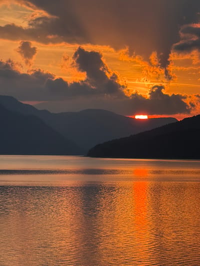 The image captures a sunset or sunrise over the Dospat Reservoir in Dospat, Bulgaria. The sky, occupying the upper portion of the frame, displays an intense gradient of orange and reddish-orange hues, with dark, fragmented clouds interspersed. The edges and some internal structures of these clouds are brightly illuminated by the sun, creating luminous orange highlights and streaks against the darker cloud bodies.

The sun is visible as a bright, narrow horizontal band of intense orange-red light, positioned just above the dark silhouette of a distant mountain range on the right side of the horizon. Multiple layers of mountains and hills stretch across the mid-ground, presented as dark, muted silhouettes in shades of deep blue-grey and black, receding into the atmospheric haze.

The lower half of the image features the surface of the reservoir, reflecting the vivid orange light from the sky. A prominent, elongated vertical streak of brilliant orange and red light, originating from the sun's position, extends across the water towards the foreground. The water's surface is textured with numerous small ripples and waves, which scatter the reflected light and create a patterned appearance. No individuals, animals, or man-made objects are discernible within the natural landscape.
