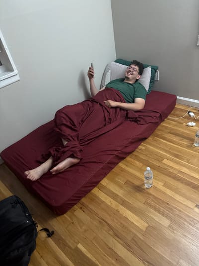 A young adult male is shown lying on a maroon-covered mattress placed directly on a light-colored hardwood floor. He is positioned diagonally across the frame, with his head resting on grey and dark green pillows against a plain light grey wall. He is wearing a dark green t-shirt and is partially covered by a maroon sheet or blanket that matches the mattress cover. The man is smiling broadly, indicating a relaxed and happy mood, and holds a silver-colored smartphone in his right hand.
The room features warm-toned hardwood flooring that occupies a large portion of the right side of the image. Against the wall behind the mattress, near the man's head, an electrical outlet with a white charger and tangled white cable is visible, along with some crumpled white paper or tissue. Two clear plastic water bottles are on the floor; one is near the top edge, and another further down to the right. A black backpack with visible zippers is situated on the floor in the bottom right corner of the frame. The setting appears to be an indoor residential space, possibly a bedroom, in Franklin Township, United States of America. The text on the water bottle labels is not legible.