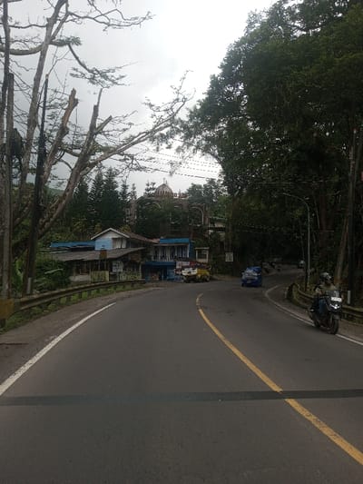 The image depicts a roadside scene in Kandapola, Sri Lanka, under an overcast sky. A two-lane asphalt road with a solid white edge line on the left and a solid yellow center line curves to the right in the mid-ground. A dark horizontal line crosses the road in the immediate foreground. On the left, a metal guardrail with a yellow section parallels the road, behind which dense vegetation, including partially bare or pruned trees, rises. Several buildings are situated on the left, behind the guardrail; one features a light blue lower section and a white upper level, while another appears to be a commercial establishment with a dark blue roof and a parked light-colored utility truck with a yellow bed in front. Further into the background, atop a hill and partially obscured by trees, stands a building with a distinctive dome structure and arched window features. On the right side of the road, lush, dark green trees form a dense canopy. A person wearing a helmet and a light-colored jacket operates a dark-colored motorcycle, moving away from the viewer on the right lane. Further down the curving road, a small blue three-wheeled vehicle is also in motion, followed by faint outlines of other distant vehicles. Utility poles and overhead wires are visible on both sides of the road.