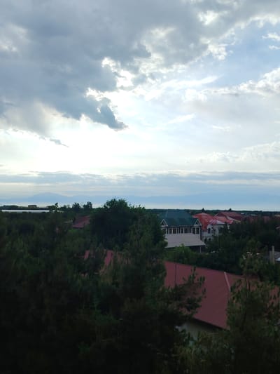 This image depicts a view of houses and trees under a cloudy sky. In the background, mountains are visible on the horizon. The foreground is dominated by dark green trees, obscuring some of the houses and their red roofs. A building with a green roof and white walls is visible in the middle ground, along with several houses with red roofs. The sky is partly cloudy with patches of bright light breaking through. The scene appears to be a residential area.