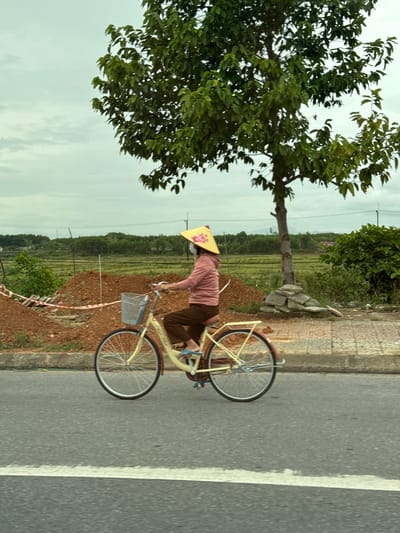 A person wearing a conical hat and a pink long-sleeved shirt is riding a cream-colored bicycle on a paved road. The person has brown pants and is wearing flip-flops. The bicycle has a silver basket attached to the handlebars and a white line is visible on the road in the foreground. Behind the person, there is a pile of reddish-brown dirt, a low green bush, and a large tree with dark green leaves. In the background, there are fields, more trees, and a cloudy sky. The scene appears to be in a rural or suburban area of Ái Tử Commune, Vietnam, suggested by the conical hat and the landscape.