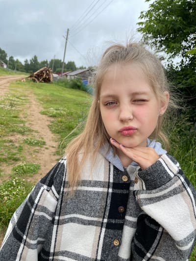 The image shows a young girl with blonde hair, wearing a plaid jacket over a grey hoodie. She is outdoors in what appears to be a rural setting, with a dirt path and green grass surrounding her. In the background, there are trees, a pile of cut wood, and some simple buildings, suggesting a village or countryside location. The sky is overcast. The girl is looking at the camera and has one eye closed in a wink, with her lips pursed. She is resting her chin on her hand. The overall scene suggests a casual, candid moment captured in a natural environment.
