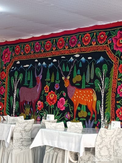 The image depicts the interior of a restaurant in Bosteri, Kyrgyzstan. A large, intricately embroidered tapestry featuring deer, trees, mountains, and flowers hangs on the wall. In the foreground, several tables are set with white tablecloths, some of which are covered with clear plastic. Chairs with ornate white covers are arranged around the tables. A table number '6' is visible on a white placard on one table, and a table number '7' on another. The ceiling is white and appears to be textured.