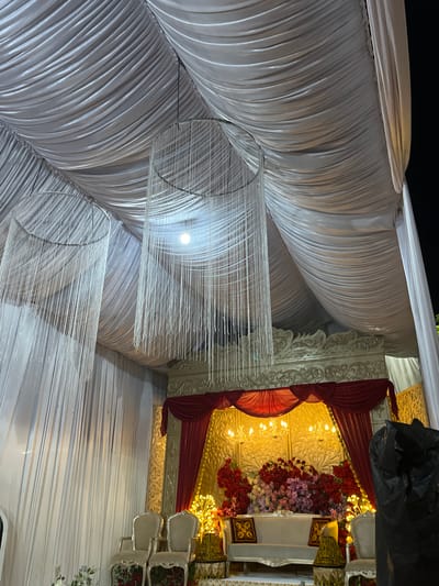 This media file captures an indoor setting decorated for an event, possibly a wedding or a festive gathering. The scene is dominated by draped white fabric, creating a soft, undulating backdrop. Suspended from the ceiling are intricate, chandelier-like decorations made of numerous fine, translucent strands, which catch the light.
In the background, a more ornate section of the decor is visible, featuring red and gold drapery, intricate white carved panels, and a lavish floral arrangement composed of red and pink flowers. Warm, yellow lights illuminate this area, suggesting it might be a stage, an altar, or a focal point for the event. A few white chairs with decorative carvings are also present, hinting at seating arrangements for guests.
The overall impression is one of elegant decoration and preparation for a celebration. The lighting and fabric suggest an evening or indoor event. There is no visible text in the image. The location context provided is Keudematangkuli, Indonesia, which could be the specific venue or region where this event is taking place.