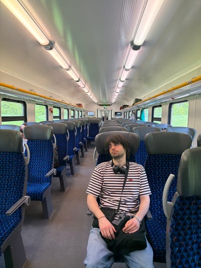 A man with dark hair and glasses lies across several seats on a train. He wears a black and white striped shirt, light blue pants, and headphones around his neck. He has a black bag on his chest. The train car has blue patterned seats and gray headrests. Windows line the side of the car, showing green foliage outside. Overhead lighting illuminates the interior. The photograph is taken from a high angle and has been rotated. There is no information available to determine the location in the city of Пески, Russia.