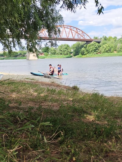 A group of four young people are on the sandy bank of a wide river, with a paddleboard partially submerged in the water. Two of the individuals are sitting on the paddleboard, while the other two are standing in the shallow water. One of the seated individuals is wearing a life vest. In the background, a rust-colored metal bridge arches over the river, with trees lining the opposite bank. The sky is partly cloudy. The foreground shows a grassy bank leading down to the water's edge. The scene appears to be a recreational outing on a river, possibly in the city of Belaya Kalitva, Russia, given the distinctive bridge structure which resembles known bridges in the area.