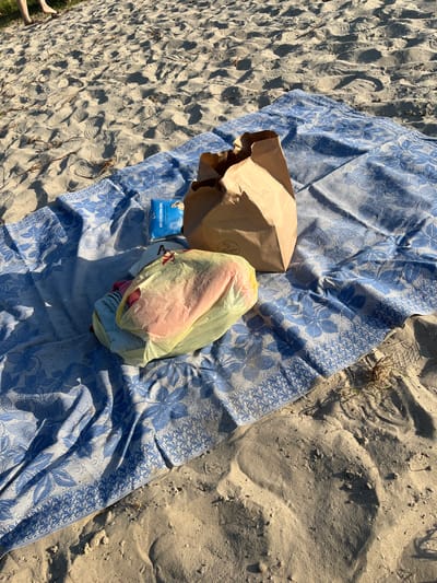 On a sandy beach in Zhurivka, Ukraine, a blue and white patterned blanket is spread out. On the blanket are a brown paper bag, a yellow plastic bag containing items that appear to be food and clothing, and a small blue package. The sandy environment is marked with footprints and some dry vegetation. In the upper left corner of the image, the legs and feet of at least two individuals are visible, suggesting they are present on the beach.