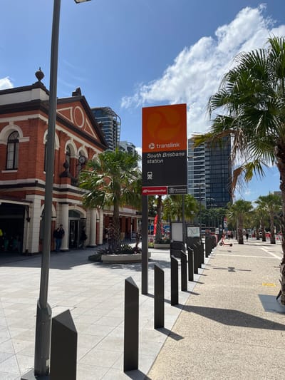 The image displays an urban street scene in South Brisbane, Australia, captured on a bright and sunny day with a clear blue sky and scattered white clouds. Strong sunlight casts elongated shadows across the light-colored paved ground.

In the mid-ground, a prominent orange and black vertical sign stands upright, clearly indicating "translink South Brisbane station." The sign also features the Translink logo, a train icon, and smaller logos for "Queensland Government" and "QueenslandRail." Below and to the left of this sign is a distinctive historic-style brick building with arched windows and a clock face, possibly an original station building. Several individuals are visible near the entrance of this building.

Further into the background and towards the right, a wide paved walkway stretches into the distance, bordered by a series of dark, angular, geometric barriers or seating elements. A lamppost is visible in the foreground on the left. Numerous palm trees are scattered throughout the scene, particularly above the station sign and lining the pathway. A modern high-rise building with dark glass facades is visible in the background. A red electric scooter is parked further down the street on the right, hinting at urban mobility. The scene depicts a lively public area, likely a station forecourt, with pedestrian activity under fair weather conditions.