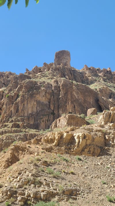The image shows a steep, rocky cliff face under a clear blue sky. The cliff is composed of layered rock formations, varying in color from light tan to dark brown. There are patches of sparse green vegetation clinging to the rock face. The cliff appears to be in an arid environment. Based on the prompt, the location is near Bostan, Pakistan. There are no people or actions visible.