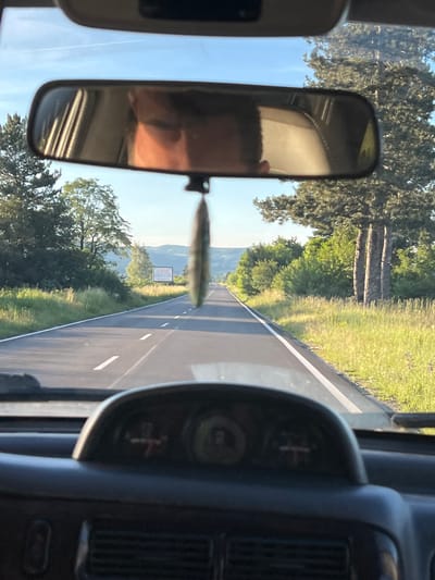The image, taken from inside a vehicle, shows a view of a two-lane asphalt road stretching into the distance under a clear blue sky with a faint contrail. The road is bordered by grassy verges and trees on either side. In the foreground, the dashboard of the vehicle is visible, with instruments including a speedometer and a fuel gauge. A rearview mirror is positioned at the top of the windshield, reflecting the interior of the car, including a person's face and part of the ceiling. A decorative item, possibly an air freshener, hangs from the rearview mirror. In the middle ground, a billboard is visible on the right side of the road. The overall impression is of a scenic drive through a rural or semi-rural area. The location is specified as Zlokuchene, Bulgaria.