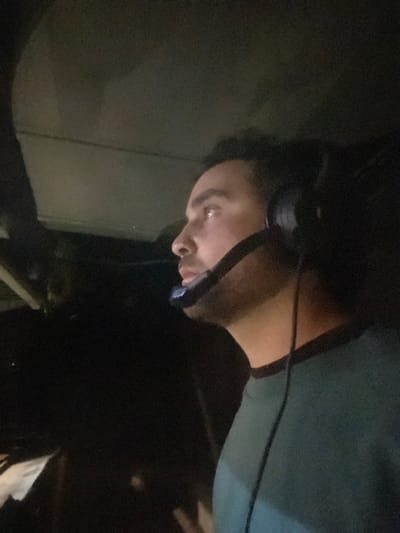 The image shows a close-up of a man wearing a headset, sitting in the driver's seat of a vehicle. He is looking to the right, appearing to be focused on something outside of the frame. The vehicle's interior is dark, with only the man and the headset illuminated. The man is wearing a teal shirt and has short, dark hair. The image was taken in Kiryat Ye'arim, Israel, at coordinates 31.806 latitude and 35.093 longitude.