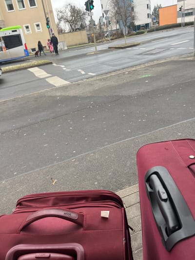 The image presents an urban street scene in Schweinfurt, Germany, from a low perspective on a sidewalk. In the immediate foreground, two maroon-colored suitcases are prominent. The upper suitcase displays a black telescopic handle with a button featuring a V-shaped or arrow-like symbol. The lower suitcase has a maroon fabric handle embossed with the text 'VIP' and a small white rectangular tag attached to its side. A cigarette butt is visible on the pavement beside the lower suitcase.

Across a wet asphalt road, a group of people, including adults and children, are seen near a crosswalk, potentially waiting to cross. A green traffic light is visible nearby. Further down the street, a white bus with green and blue accents is present. Buildings and bare trees line the street in the background. The overcast sky and damp road suggest a cloudy day, possibly after rain.