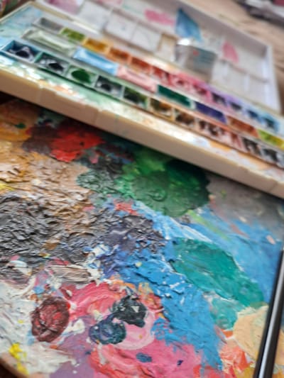 This media file captures a close-up view of art supplies. The primary subjects are a watercolor paint palette with many individual pans of color, and a larger palette covered in thick, wet paint in various vibrant hues, including blue, green, pink, red, and yellow. The paint palette shows signs of recent use, with textured swirls and dollops of paint. A dark, thin object, likely a paintbrush, is visible resting across the top of the paint palette, its tip possibly touching the paint.

The scene appears to be an artist's workspace, with the watercolor palette laid out to the left and the mixing palette to the right. The lighting suggests an indoor setting, possibly a studio or a home environment. There are no people visible, and the focus is entirely on the artistic tools and materials. The colors are rich and saturated, indicating a lively and creative atmosphere.

Notable details include the textural quality of the paint on the mixing palette, which is thick and impasto-like. The watercolor pans are neatly arranged, contrasting with the more chaotic application of paint on the larger palette. The overall impression is one of an artist actively engaged in their craft.

No visible text is present in the image. Given the location context of Popovo, Bulgaria, the image depicts typical art supplies that might be found in such a setting.