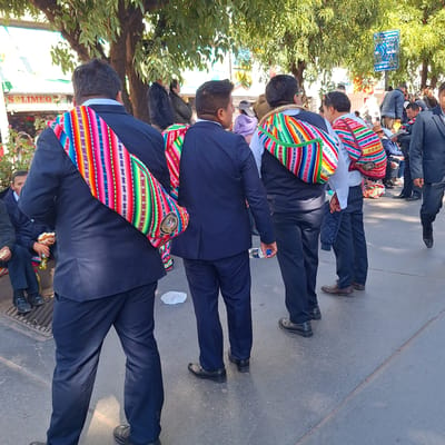 The image depicts a street scene in Wanchaq, Peru, with several men wearing dark suits and colorful woven sashes draped over their shoulders. These sashes feature intricate patterns and vibrant colors, including red, pink, yellow, green, blue, and white stripes, as well as geometric motifs. The men appear to be standing or walking on a paved street, with trees providing shade overhead. In the background, there are buildings and other people. One man in the foreground is holding a bottle of water. Another man is seen reading from a document. A blue sign with white lettering and an image is visible on a pole in the background. The overall impression is of a cultural or celebratory gathering.