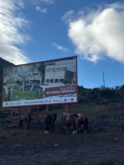 The image displays an outdoor scene in San Vicente Pacaya, Guatemala, dominated by a large billboard and a group of horses.
A prominent rectangular billboard, framed in red and supported by red poles, occupies the midground. It advertises "Santa Teresita Hotel & Spa Termal" and "Enjoy the Best HOT SPRINGS EXPERIENCE," featuring a photographic depiction of a stone building with multiple windows and terraced natural pools surrounded by palm trees. The billboard also displays contact information: a phone number (6644 1000), a website (www.santateresita.com.gt), and social media icons (WhatsApp, Facebook, Instagram, TikTok).
Below the billboard, seven horses are present. They are equipped with saddles and bridles, indicating they are prepared for riding. Their coats vary in color, including shades of brown, dark brown, and dappled grey. The horses are standing idly on dark, uneven ground composed of earth or volcanic ash.
The background consists of a steep hillside covered with sparse, dark green vegetation. The sky above is clear blue with scattered white, wispy clouds. Thin, dark lines, possibly power lines, cross the upper sky, with a vertical pole visible on the right side. No human figures are visible in the scene. The setting appears to be a staging area or rest stop in a rugged, natural environment.