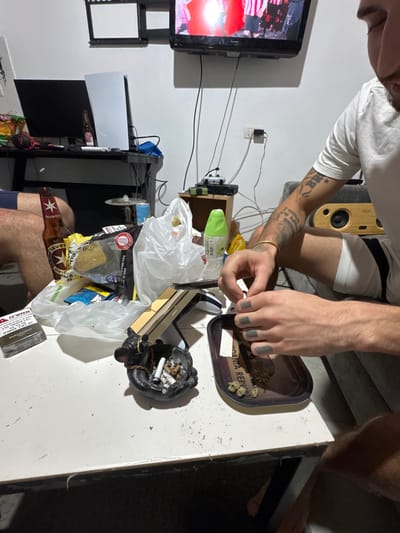 This image depicts an indoor scene, likely a living room or recreational area. A specific city within Gilboa Regional Council, Israel, is not discernible from the image, though an Israeli beer brand is present on the table.
On the right, a person with tattoos on their right arm and grey-painted fingernails/knuckles is actively rolling a substance using a metal tray labeled "California Republic," which contains green plant matter resembling cannabis and some smaller buds. Rolling papers are visible near the tray. A black ashtray filled with cigarette butts, ash, and a golden ring rests centrally on a white rectangular table. To its left is a pack of Parliament cigarettes. A crumpled clear plastic bag containing various snacks, including chips, is also on the table, alongside a green plastic bottle and a brown beer bottle with a star emblem, consistent with an Israeli brand (Maccabee). Small crumbs and debris are scattered across the table surface. The legs of a second individual, wearing blue shorts, are partially visible on the far left.
In the background, a white wall features a wall-mounted black flat-screen television displaying content with prominent red and white colors. Below the television, a white PlayStation 5 console and a black monitor are situated on a black desk. Numerous cables and wires are visible running down the wall and behind the desk. To the right of the desk, a wooden speaker or radio unit is visible on a grey surface. The room appears to be illuminated by artificial light and has a somewhat cluttered appearance.