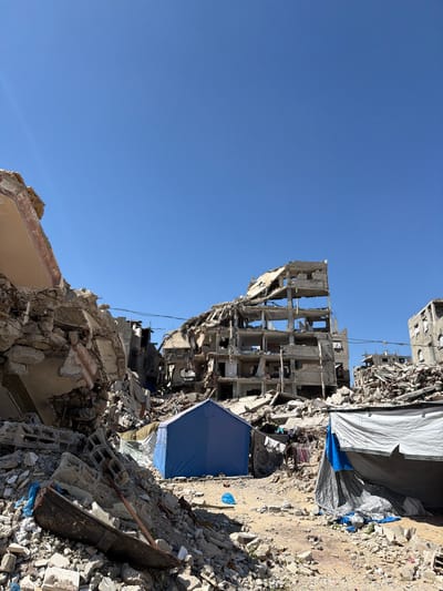 The image depicts a scene of widespread structural devastation in Jabalia, Palestinian Territory. Under a clear, bright blue sky, the foreground and mid-ground are covered in extensive rubble piles composed of fractured concrete, rebar, and fragmented building materials. In the central background, a multi-story concrete building, estimated at five to seven floors, exhibits severe structural damage, with upper sections collapsed, exterior walls largely absent, and internal framework exposed. Additional partially destroyed buildings and large debris mounds are visible in the background and to the right.
In the mid-ground, a blue, approximately rectangular tent is erected directly on the rubble-strewn ground. To its right, a larger, improvised shelter constructed from white and blue tarpaulins is present, with articles of clothing hung for drying on a line nearby. To the left of the blue tent, a dark, trough-like object is partially submerged in debris. A small, blue plastic bag lies on the ground near the blue tent. No individuals are visible. Utility lines span between damaged structures in the deeper background.