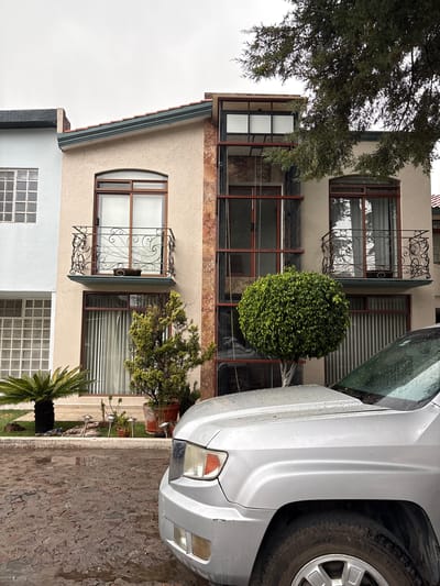 A multi-story building in San Francisco Totimehuacán, Mexico, exhibits a light beige facade with brown window frames. The windows on the upper floors feature ornate wrought iron decorative elements. A green, spherical bush is positioned on one of the floors. The front of a silver car is prominently visible on the right side of the image. A stone wall borders the property. A small palm tree is visible in the front yard. Overcast sky conditions.
