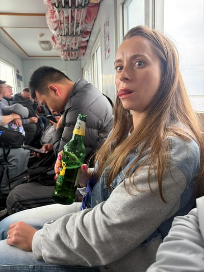 An eye-level shot captures a woman looking playfully at the camera, her tongue slightly out, inside what appears to be a passenger cabin of a ferry or similar public transport. She has long, light brown hair and is wearing a light gray sweatshirt over a denim jacket, paired with light-colored jeans. In her right hand, adorned with bright pink and orange nail polish, she holds a green bottle, likely beer, with a prominent yellow label displaying Asian characters. A gold ring is visible on her left ring finger.
Behind her, a man in a dark puffer jacket is partially visible, facing away from the camera. Several other passengers are also visible, seated or standing, filling the compartment. The setting features rows of shelves overhead, stacked with numerous red-packaged items, possibly emergency equipment. Daylight streams through windows along the side of the cabin. Some red symbols or text are visible on a white panel beneath a window. The overall scene conveys a casual travel environment during the day.