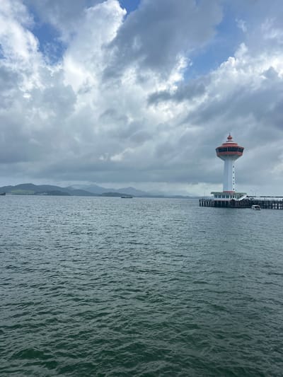 An overcast day is depicted in this image, with the sky filled with a mixture of white and gray clouds. In the mid-ground, a tall white lighthouse stands on a pier, its top section a vibrant red. The lighthouse has a modern, multi-story observation deck with numerous windows. The pier extends into the dark, choppy water, and a small white boat is docked on the right side. In the background, a hazy coastline with rolling hills and mountains is visible. The water in the foreground is a deep, dark green, with visible ripples. The scene appears to be a coastal area, likely a port or harbor. The location is identified as Ban Hin Chang, Thailand.