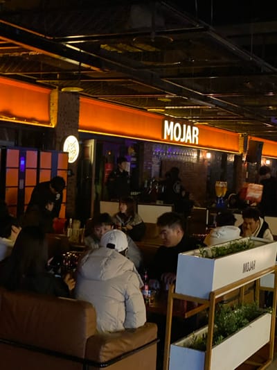 The image captures a vibrant night scene inside or at the entrance of a bustling establishment, likely a bar or restaurant, illuminated primarily by artificial lights. The photograph is oriented vertically but appears to have been taken sideways, requiring a 90-degree clockwise rotation to view it correctly.
In the upper portion of the image, a large, prominent sign featuring the word "MOJAR" in bright, white, capital letters against a glowing orange background stretches horizontally across a wall. This sign is a central source of light, casting a warm orange hue on its immediate surroundings. Below this main sign, the interior space is filled with people seated at tables and couches, engaged in various activities.
To the right side of the frame, there's a comfortable seating area with brown leather couches or booths. Several patrons are visible here. One person in the foreground, wearing a light-colored (possibly white or light grey) puffer jacket and a white baseball cap, is seated and appears to be looking towards the left side of the scene. Other individuals are seated around them, some facing away, others seemingly interacting. White, rectangular planter boxes filled with green foliage act as decorative dividers within this seating area. One of these planters also clearly displays the word "MOJAR" in black text on its side.
Towards the mid-ground and left of the seating area, more people are scattered throughout the establishment. Some are at smaller tables, while others are standing near what appears to be a bar or service counter, indicated by rows of glasses. The lighting in these areas is dimmer, relying on overhead lights or spotlights, creating a cozy evening ambiance. Below the large "MOJAR" sign, there's a dark metal railing, suggesting an elevated section or balcony. Further down, another smaller, circular illuminated sign with an indistinguishable logo or text is visible. Below this, a decorative wall segment composed of light-colored bricks or tiles features embedded blue glowing elements, adding to the modern aesthetic.
The overall atmosphere appears relaxed and social, with people enjoying their evening out. The time of day is clearly night, as evidenced by the dark background and the reliance on interior and sign lighting. No specific weather conditions are discernible, but the presence of puffer jackets suggests it might be cool weather.