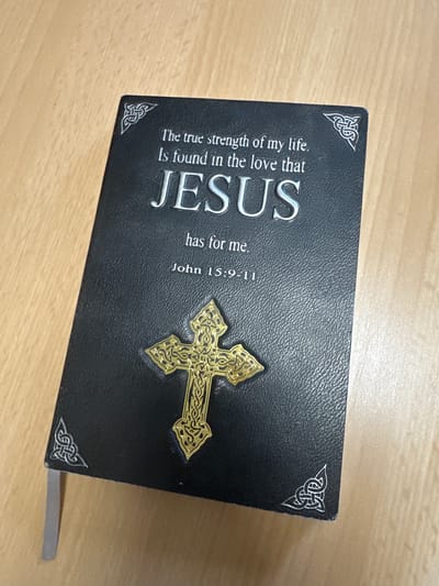 A close-up, high-angle shot captures a black book with silver Celtic knot designs in each corner. The book is open to a page featuring text and a golden, ornate cross emblem. The text reads: "The true strength of my life. Is found in the love that JESUS has for me. John 15:9-11". The book is resting on a light-colored wooden surface. There is no indication of the city of Bad Blankenburg, Germany in the image.