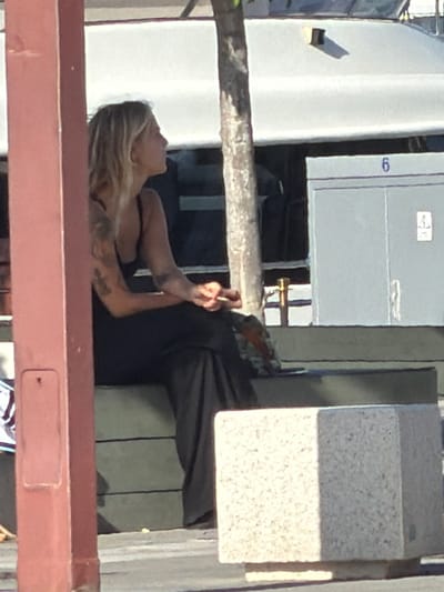 In Eivissa, Spain, a blonde woman with tattoos on her arms sits on a bench, looking to her right. She wears a black top and pants. Her right hand is raised, holding a cigarette to her mouth, as if she is smoking. Behind her, a tree trunk is visible, and to the right, a light gray structure with the number '6' in blue is mounted on a building. The scene appears to be outdoors, with a paved surface in the foreground.