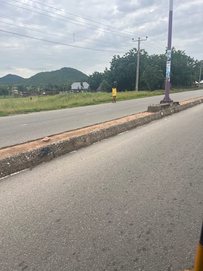 A person wearing a yellow shirt and black shorts stands on the side of a paved road in Kona Garu, Nigeria. The person is facing away from the camera. The road is bordered by a grassy shoulder and a concrete curb. In the background, there are trees and a mountain. Overhead power lines are visible. The sky is overcast, suggesting it might be a cloudy day. A purple and white pole with posters attached to it stands near the side of the road. The posters appear to contain images of people and text, possibly political campaign material given the location context. The scene appears to be set during the daytime.