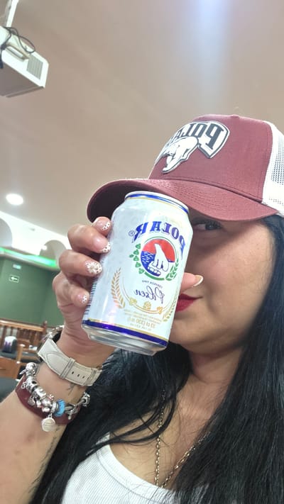 A woman, partially visible, holds a can of "Polar" beer to her lips while wearing a maroon and white trucker hat. Her nails are decorated with a light-colored polish and small, scattered white dots, and she has red lipstick on. She is also wearing a white watch on her left wrist and a multi-strand bracelet with various charms. The background shows a ceiling-mounted projector and a green wall with a fire exit sign. The setting appears to be an indoor public space, possibly a bar or restaurant, located in Torrejón de Ardoz, Spain.
