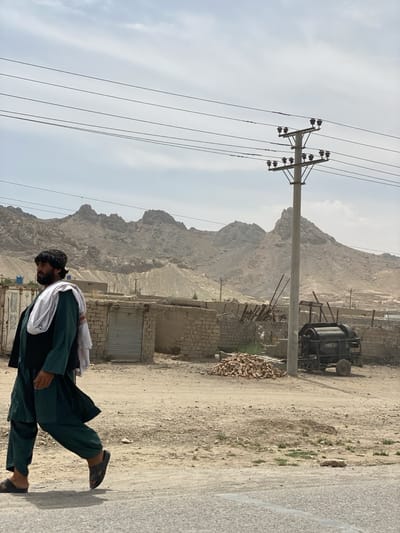 The image portrays a dusty, arid landscape in Dand, Afghanistan, under an overcast or hazy sky. A man in traditional attire walks from left to right across the mid-ground. He wears a dark green shalwar kameez, a black vest, and a white shawl draped over his left shoulder, completing his look with dark sandals. His expression is not clearly visible as he moves along a transition from a paved road in the foreground to a dirt path.
In the middle ground, simple, low-rise buildings with light-colored brick or mud walls are visible, some featuring corrugated metal doors. To the far left, a portion of a blue shipping container can be seen. Towards the center-right, a pile of reddish-brown bricks sits on the ground. Further right, a dark, wheeled piece of agricultural machinery, resembling a thresher, is parked.
A prominent concrete utility pole with multiple insulators supports several power lines stretching across the upper part of the frame, bisecting the view of the distant background. The horizon is dominated by rugged, barren mountains with rocky peaks, characteristic of the region. The scene depicts a daytime setting, likely late morning or afternoon, given the bright yet diffused light, in a rural or semi-urban environment defined by its dry climate and mountainous terrain. No visible text is present in the image.