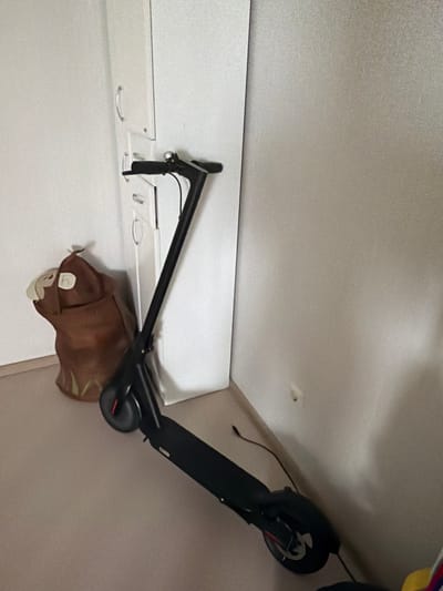 The image depicts an interior scene, likely a residential or storage area, where a black electric scooter is propped upright against a white cabinet. To the left of the scooter, a brown, bag-like object with a cartoonish face is visible. The background consists of a plain, light-colored wall. The scene is devoid of any human presence or overt activity. The overall impression is one of stillness and storage. The question about Crokhovo, Russia appears to be a misinterpretation as there is no visual information within the image to suggest this specific location.