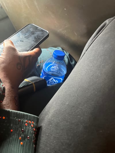 Within a vehicle in Shuwarin, Nigeria, the right hand of an individual is positioned centrally, holding a dark-colored smartphone with a dark, reflective screen. The hand exhibits intricate dark temporary tattoo patterns on the dorsal surface and thumb. The individual wears a dark garment, possibly a sleeve, accented with small, reflective red beads or embellishments. Adjacent to the hand and resting on dark, ribbed fabric is a clear plastic bottle of "Fresh" brand water with a blue cap and a blue label featuring a mountain logo. A dark, rectangular bundle, appearing to be currency or documents, is secured with a light-colored elastic band and rests between the hand and the water bottle. The background consists of a dark, textured interior surface, suggesting a vehicle compartment.
