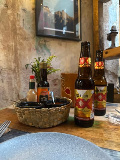 The image depicts a table setting at a restaurant in Zacatecas City, Mexico. On a rustic wooden table, two bottles of Dos Equis Amber Especial beer stand prominently. Beside them, a woven basket holds two small black dipping bowls, one containing a reddish sauce, and a bottle of what appears to be hot sauce. A bottle of Crosse & Blackwell Worcestershire sauce is also present. In the background, a framed artwork hangs on a textured wall, and part of a terracotta pitcher is visible. The overall ambiance suggests a casual dining experience.