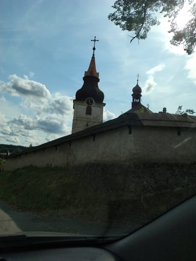 This is a photograph taken from a vehicle in Hlávkov, Czechia, looking towards a fortified church complex. The main subject is a tall, white church tower with a dark, curved roof and a smaller, onion-domed structure next to it, also topped with a cross. A long, weathered stone wall surrounds the complex, with a dark, sloping roofline visible along its top. Lush green grass grows on the slope leading up to the wall, and a tree branch with leaves is visible in the upper right corner, silhouetted against a bright, cloudy sky. The sun is shining, creating lens flare in the upper right quadrant of the image. The foreground shows the interior of a vehicle, with the dashboard and windshield wipers visible.