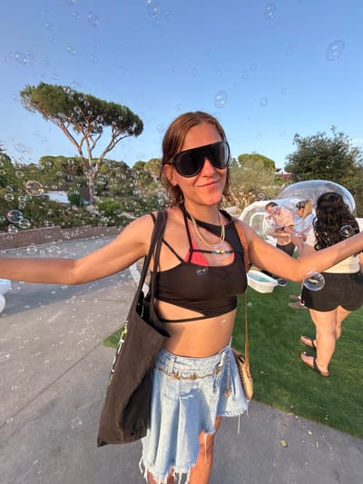 A young woman with brown hair and sunglasses is standing outdoors, possibly at a park or event in Sant Vicenç De Montalt, Spain, as indicated by the surrounding vegetation and architecture. She is wearing a black crop top with a pink bikini underneath, a light blue denim skirt, and a gold chain necklace. She holds a black tote bag over her shoulder. In the background, other people are visible near a large inflatable bubble structure, and many bubbles are floating in the air, suggesting a festive atmosphere. The scene is illuminated by natural light, likely from the sun.