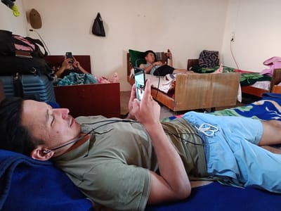 In a room in Gulshan, Uzbekistan, three individuals are seen lying on beds, each engrossed in their mobile phones. The primary subject, in the foreground, is a man lying on his left side on a blue blanket. He is wearing a green t-shirt and light blue shorts, with earbuds in his ears. His right hand is holding a smartphone, and his gaze is directed towards it. To his left, luggage, including a suitcase and bags, is stacked near a wooden bed frame. In the background, two other individuals are also lying on beds, each similarly occupied with their phones. One person is on a bed with a green pillow, wearing a striped shirt and dark shorts, holding a phone. The other person is on a bed further back, with their feet visible, also using a phone. The room appears to be a dormitory or a shared living space, with multiple beds and sparse wall decorations, including a hanging bag and a brown hat.