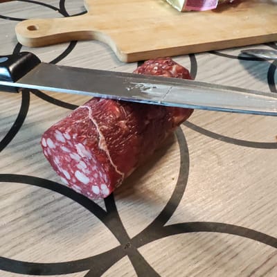 A close-up, eye-level shot captures a salami being sliced with a large kitchen knife. The salami, characterized by its dark red meat and white fatty marbling, is partially cut, revealing its cross-section. The knife's metallic blade is positioned horizontally across the salami, with its handle visible on the left. In the background, a light-colored wooden cutting board and a yellow object, possibly butter or cheese, are present. The scene is set on a table with a light wood grain surface adorned with a black, abstract circular pattern. The lighting is natural and diffused, casting soft shadows. The image does not depict any specific location in Isakovo, Russia; it appears to be a domestic setting.