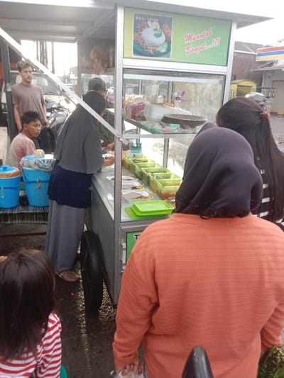 People are gathered around a food cart selling "Nasi Uduk." A woman in a gray hijab is serving customers from behind the cart, which has food containers and plates on display. Customers are visible in front of the cart, including a person in an orange sweater with a hood and a child wearing a striped shirt. Other customers stand to the side, waiting for their turn to order. Buckets are on the left side of the cart. A tire is visible on the cart's side, suggesting it's mobile.