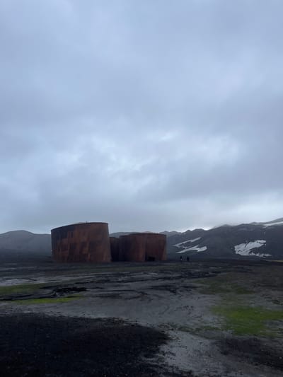The image shows rusty, cylindrical structures in a row along a dark, cracked ground with patches of green vegetation. In the distance, mountains with snow patches rise behind the structures, all under a cloudy sky. Two small figures stand further along the line of cylinders, giving a sense of scale in the vast landscape. The overall impression is bleak and industrial within a rugged natural environment.
