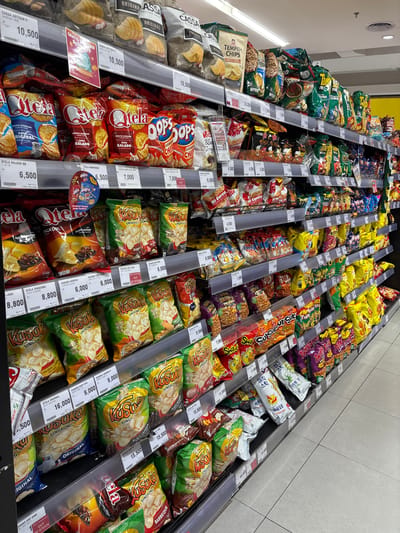 The image captures a vibrant, well-stocked snack aisle inside a supermarket or grocery store in Kelapa Dua, Indonesia. The scene is dominated by numerous bags of various chips, crackers, and instant noodles displayed in long, horizontal bins or shelves, creating an organized yet abundant arrangement. No people are visible, focusing entirely on the products.

The activity depicted is the merchandising and display of snack food items for sale. Notable details include the clear pricing labels, predominantly in Indonesian Rupiah (e.g., 10,500, 6,500, 7,000, 8,800, 6,000, 15,500), attached to the edges of the display units. Several "OUT OF STOCK" signs indicate popular or temporarily unavailable items. The bright, indoor lighting suggests an active retail environment, though the exact time of day is not discernible. The products are largely pre-packaged in colorful, branded plastic bags.

Visible text on the packaging and labels includes brand names such as CASSA, TEMPEH CHIPS, QTELA, OOPS, KUSUKA, Mi Goreng, and SuperSuSu. Product descriptions and flavors are also prominent, like "ARTISAN ORIGINAL FLAVOR," "KERIPIK SINGKONG RASA BALADO" (cassava chips with balado flavor), "CHICKEN CURRY," "BLACK PEPPER," "BARBEQUE," and "LADA HITAM" (black pepper). Some packaging also features Korean text such as "매콤달콤" (spicy and sweet) and "매직 스틱" (magic stick), indicating a mix of local and potentially imported products. A "GRATIS RP" (Free Rupiah) sign and a "New" label for "OOPS" chips are also seen. The overall impression is one of a diverse selection of snacks catering to various tastes.